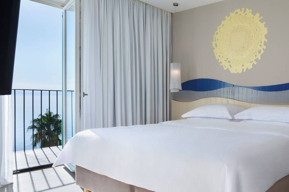 A hotel room with a bed, a view of the sea, a curtain, a wall decoration, and a nightstand.