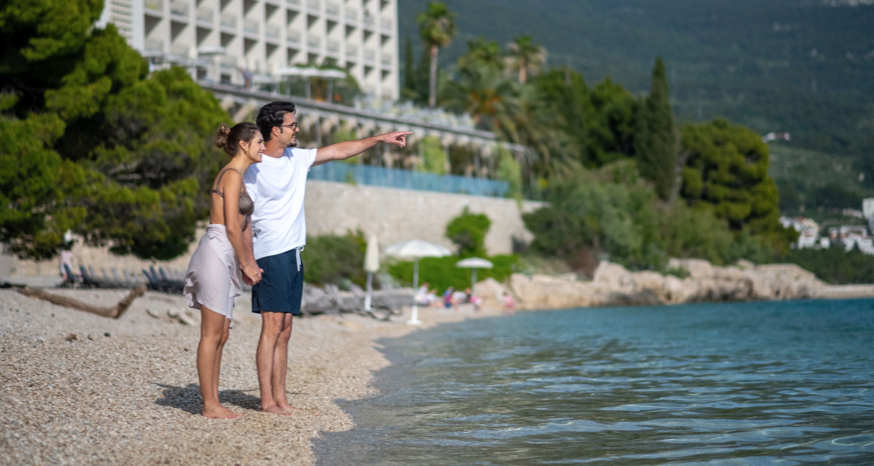 A couple standing on a pebbled beach with a view of a building and greenery in the background.