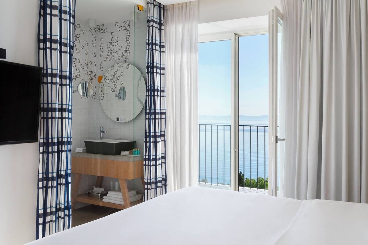 A hotel room with a large bed, a bathroom with a sink and mirror, and a balcony with a view of the sea.