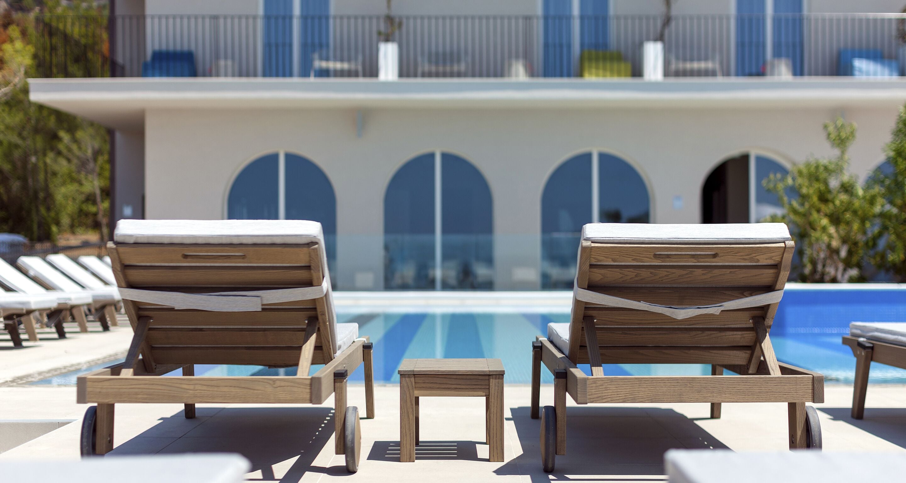A poolside area with wooden lounge chairs and a small table, overlooking a pool with a modern building in the background.