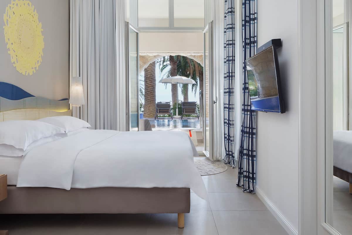 A modern hotel room with a bed, a bedside table, a TV mounted on the wall, and a view of the outdoor area.