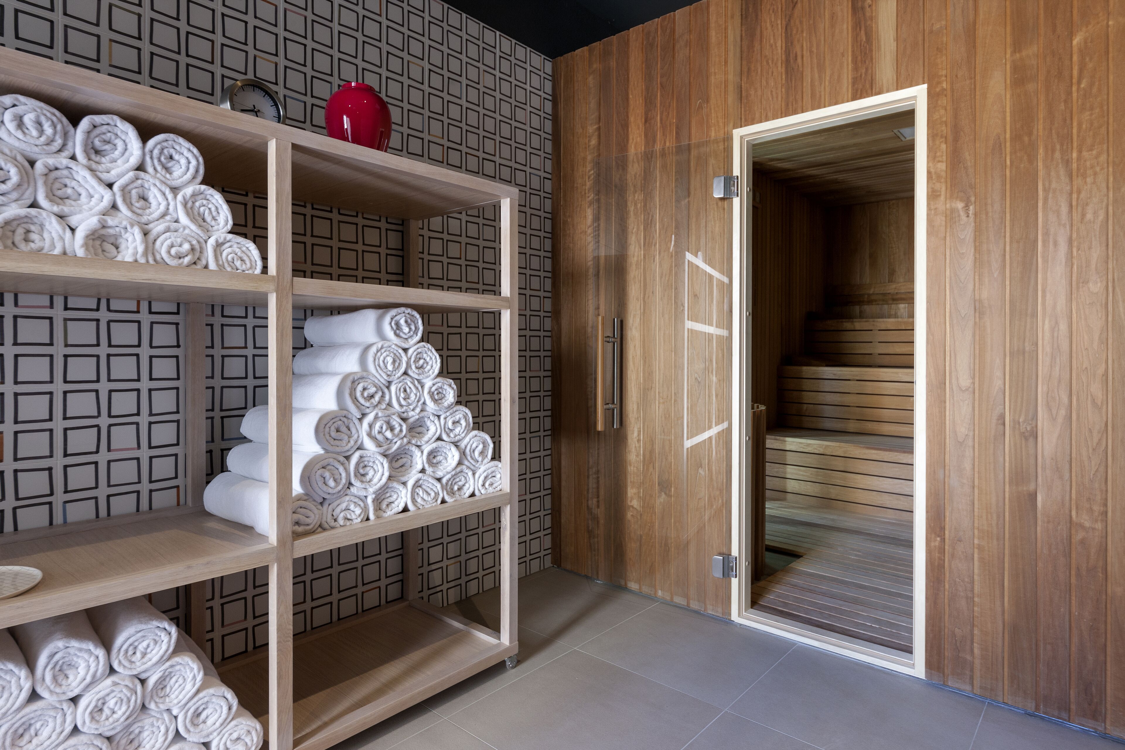A modern bathroom with wooden shelves holding rolled towels, a red vase, and a wooden door leading to a sauna area.