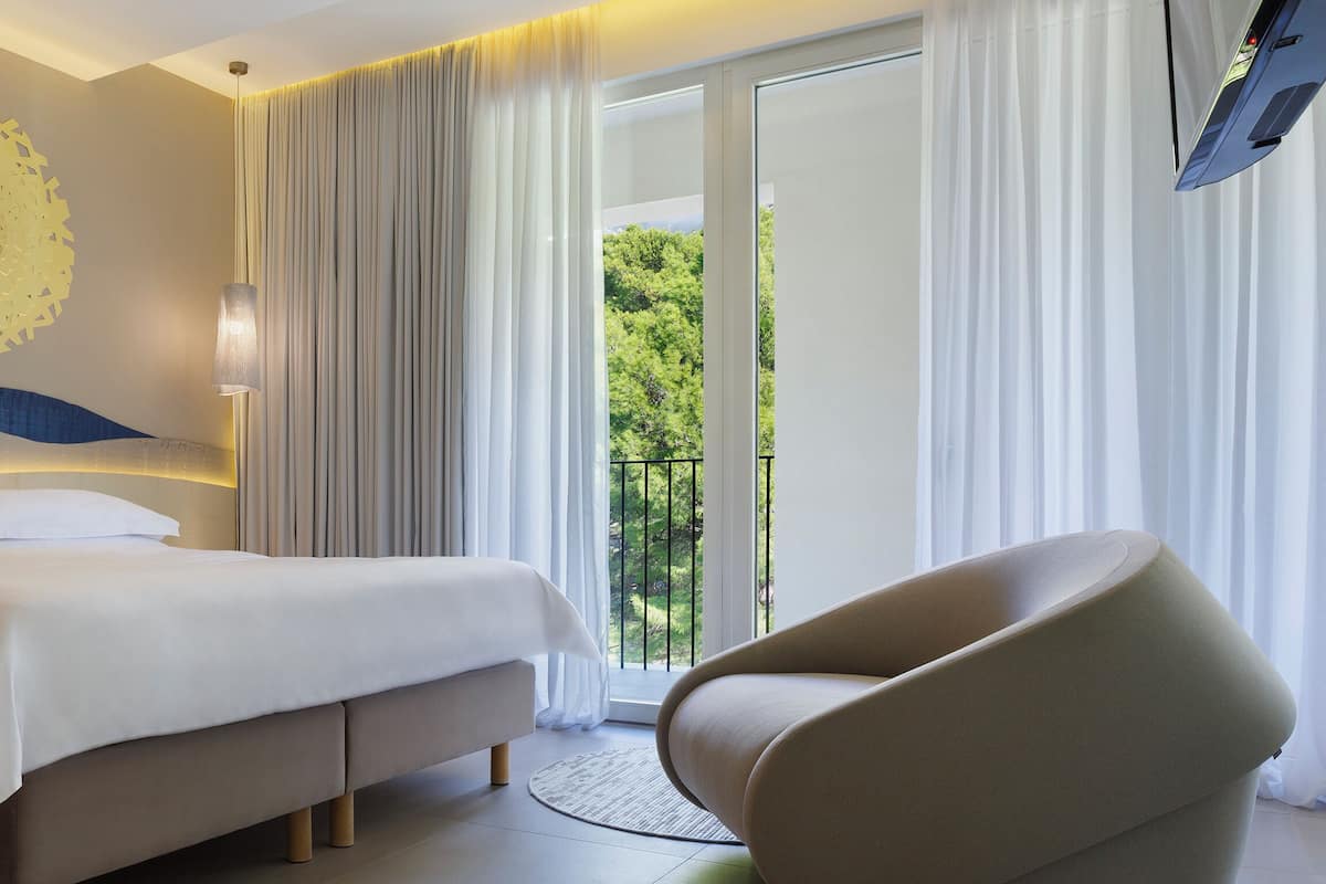 A modern hotel room with a large bed, a comfortable armchair, a wall art piece, and a balcony with a view of greenery.