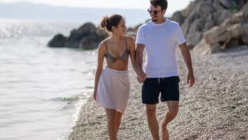 Una pareja caminando sobre una playa de guijarros con un litoral rocoso y un mar tranquilo al fondo.