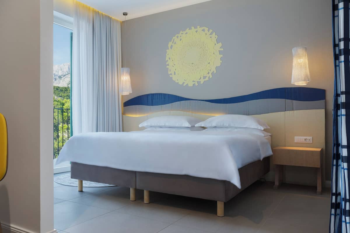 A modern hotel room with a bed, a chair, a bedside table, and a decorative wall piece.