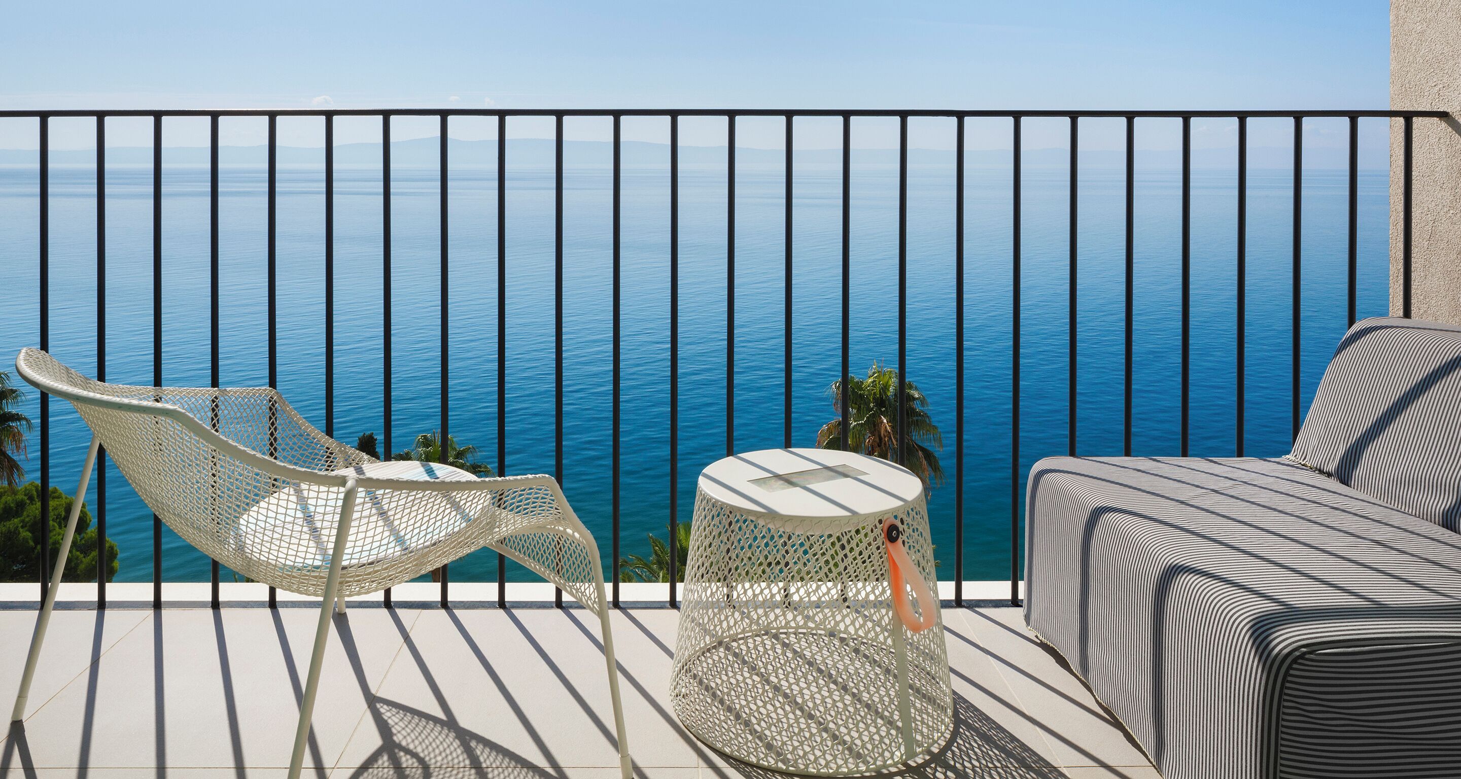 Ein Balkon mit weißem Stuhl, einem kleinen runden Tisch und einer Couch mit gestreifter Sitzkissen, mit Blick auf das ruhige Meer.