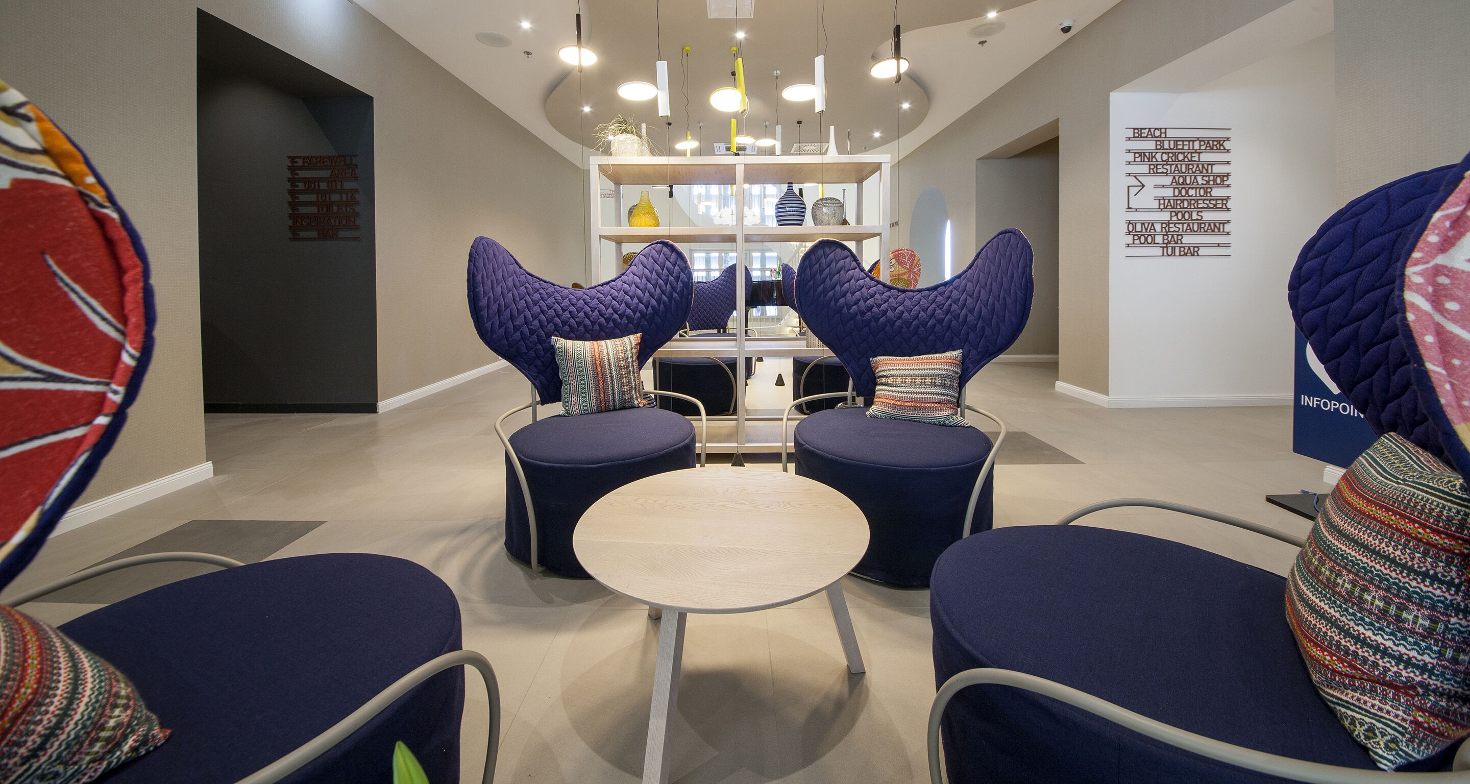 Un espace de détente moderne avec des tables rondes et des fauteuils bleus rembourrés, une étagère blanche en arrière-plan et un mur indiquant les équipements disponibles, comme une plage et un bar au bord de la piscine.