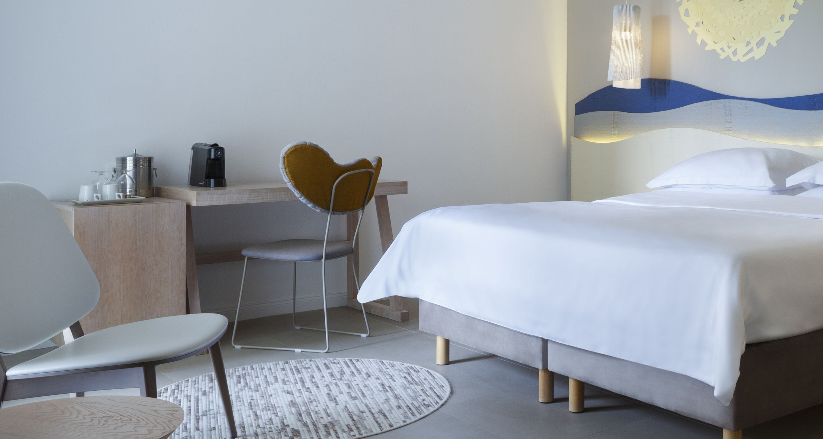 Una camera d'albergo con un letto, una scrivania con una macchina per il caffè, una sedia e un elemento decorativo a parete.