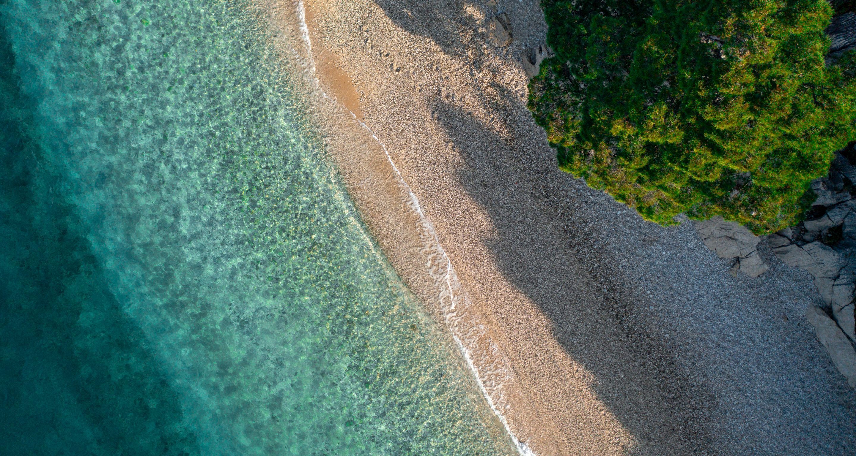 An aerial view of a beach with clear turquoise water, a sandy shore, and green vegetation on the right.