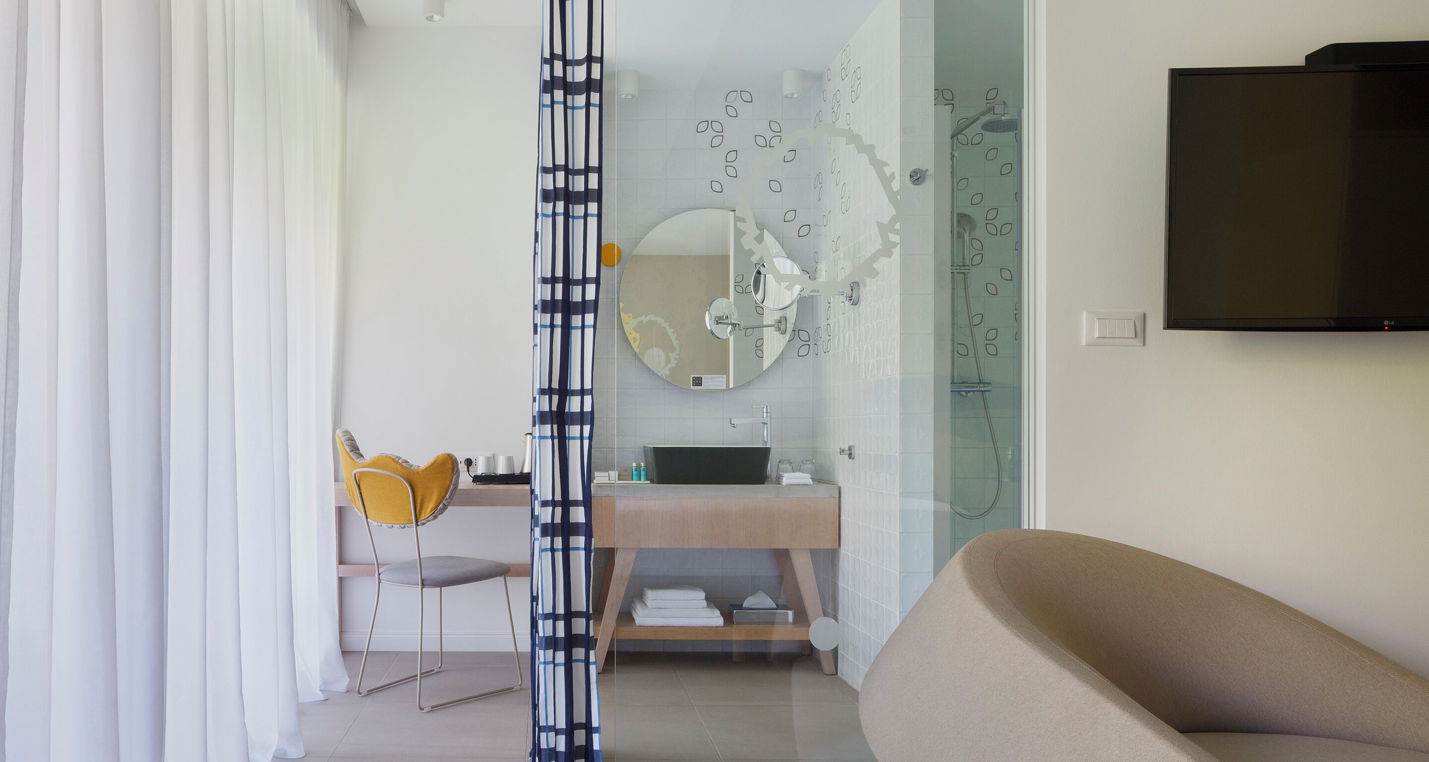 A modern bathroom with a shower, a sink, and a mirror. There is a TV mounted on the wall and a chair with a yellow cushion.