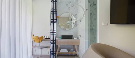 A modern bathroom with a shower, a sink, and a mirror. There is a TV mounted on the wall and a chair with a yellow cushion.
