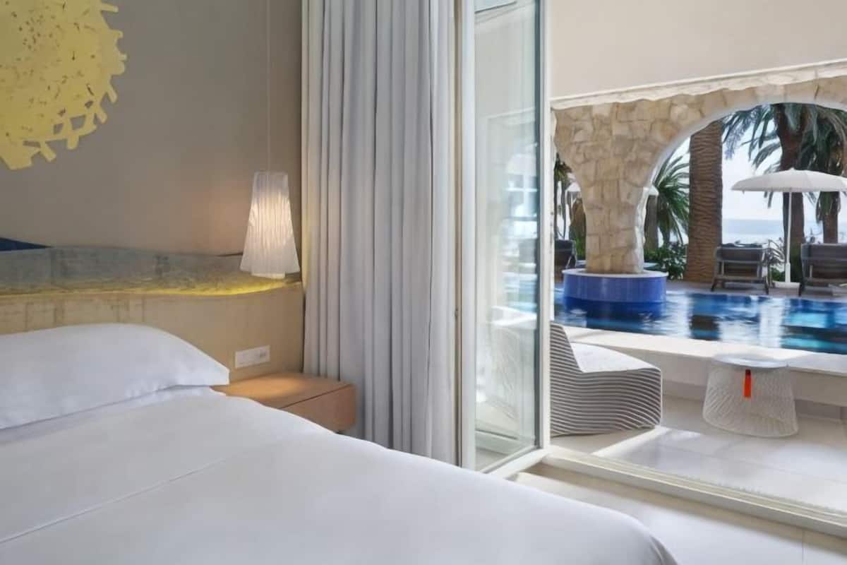 A hotel room with a bed, a nightstand, a lamp, a glass door leading to a pool area with a view of the ocean.