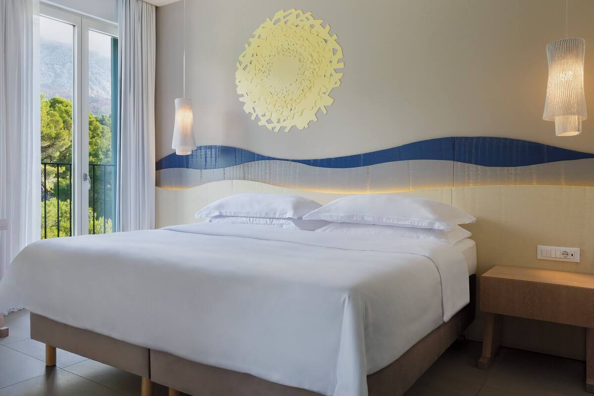 A hotel room with a bed, a chair, a nightstand, and a view of greenery outside.