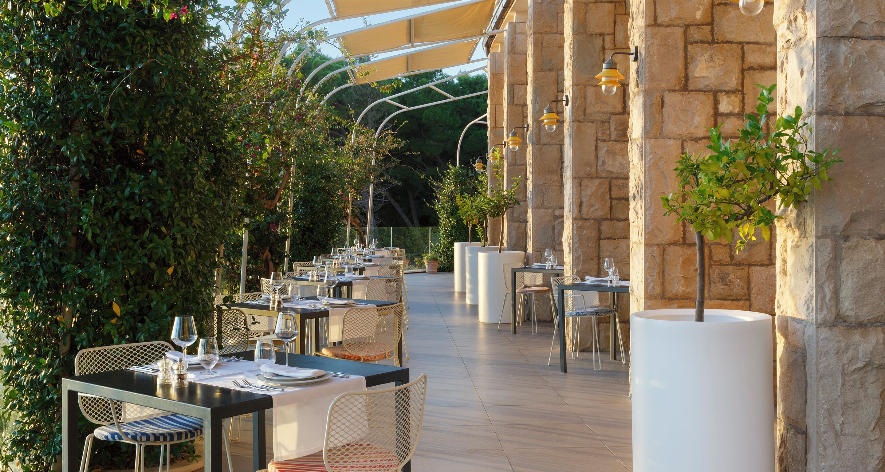 An outdoor dining area with tables and chairs, surrounded by greenery and stone walls.