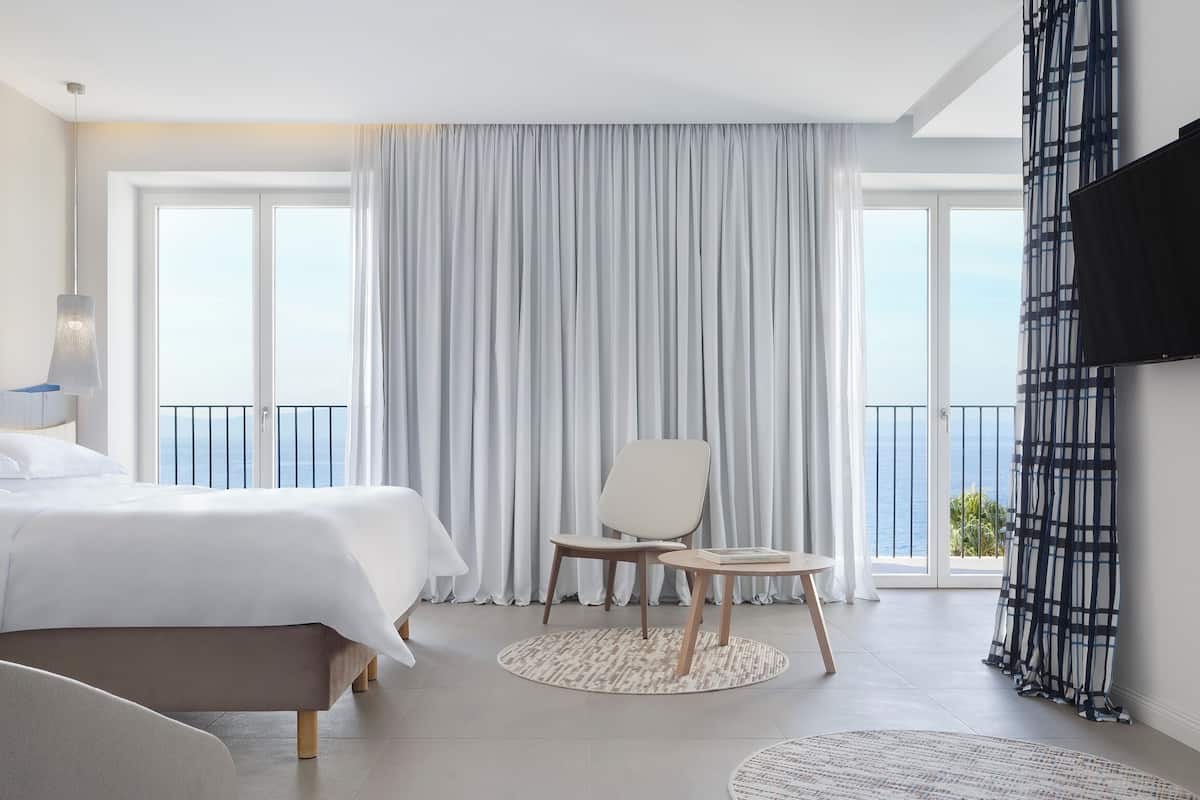 A modern hotel room with a large bed, a chair, a small table, and a view of the sea through large windows.