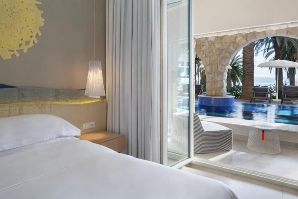 A hotel room with a bed, a nightstand, a lamp, a glass door leading to a pool area with a view of the ocean.