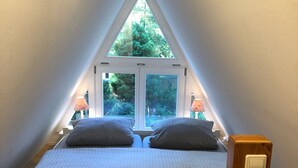 A bedroom with a sloped ceiling, a bed with a headboard, two bedside lamps, and a window with a view of trees.