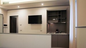Ein modernes Hotelzimmer mit einem Bett, einer kleinen Küchenzeile, einem Flachbildfernseher und einer Tür zu einem weiteren Raum.