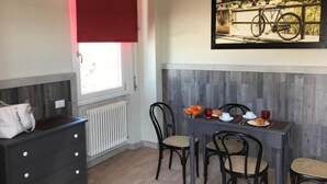 Een eethoek met een tafel gedekt voor vier personen, een radiator, een raam met een rode zonwering, en een lijst met een afbeelding aan de muur.