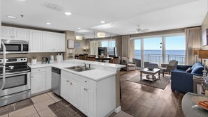 A modern kitchen with white cabinetry and stainless steel appliances, a dining area with a view of the ocean, and a living room with a blue sofa and a large window. - Gulf Views~Steps2Beach~FAB Lagoon Pool~2BR Sterling Shores 916 (Destin)