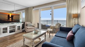 A spacious living area with a large sofa, a coffee table, and a television displaying a sunset. There is a balcony with a view of the ocean. - Gulf Views~Steps2Beach~FAB Lagoon Pool~2BR Sterling Shores 916 (Destin)