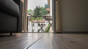 A room with a wooden floor, a sofa, and a view through a glass door to an outdoor balcony with a railing. - Bilocale Mameli - With balcony (Verona)