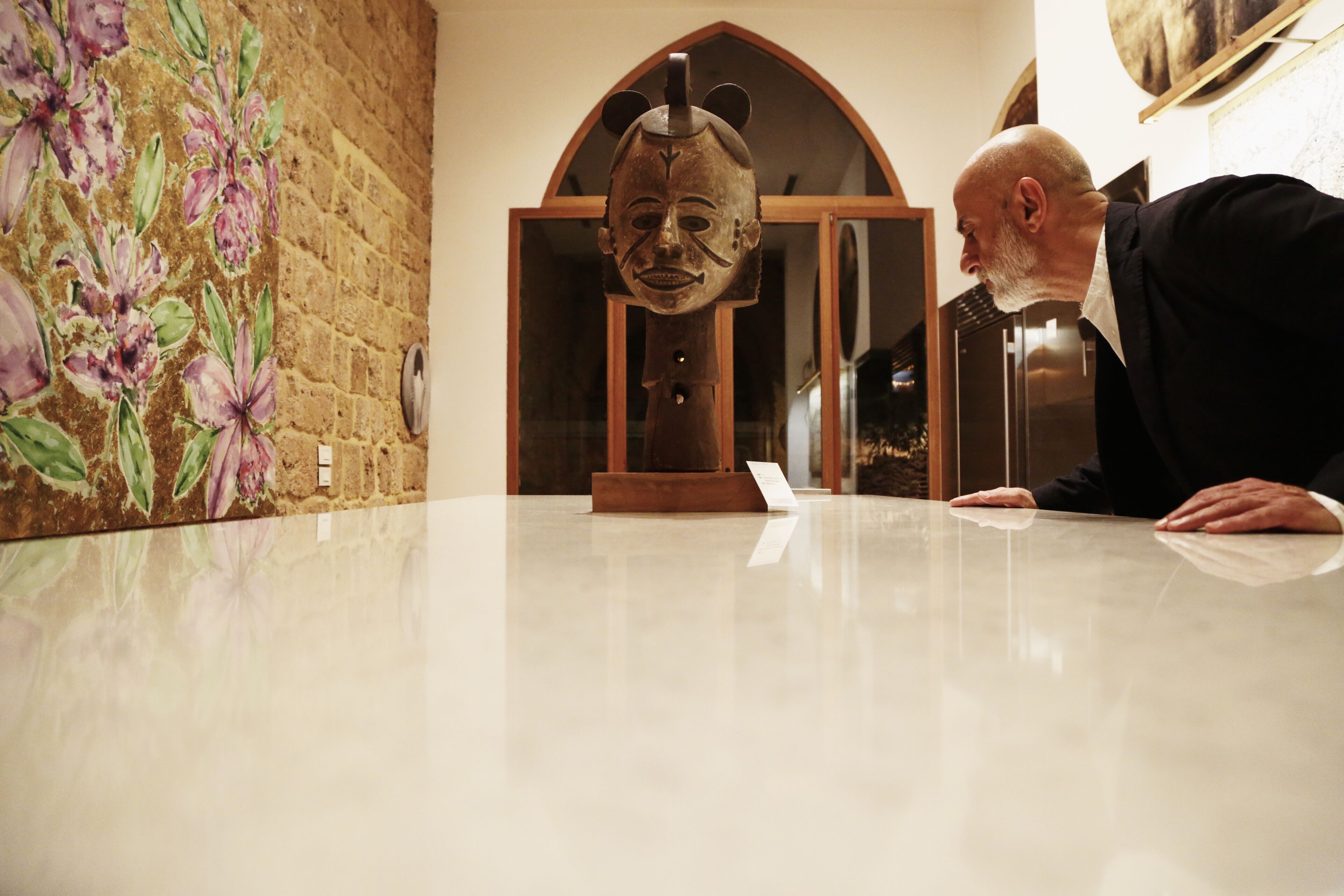 A man leaning on a marble table in a room with a floral mural and a wooden arched doorway.