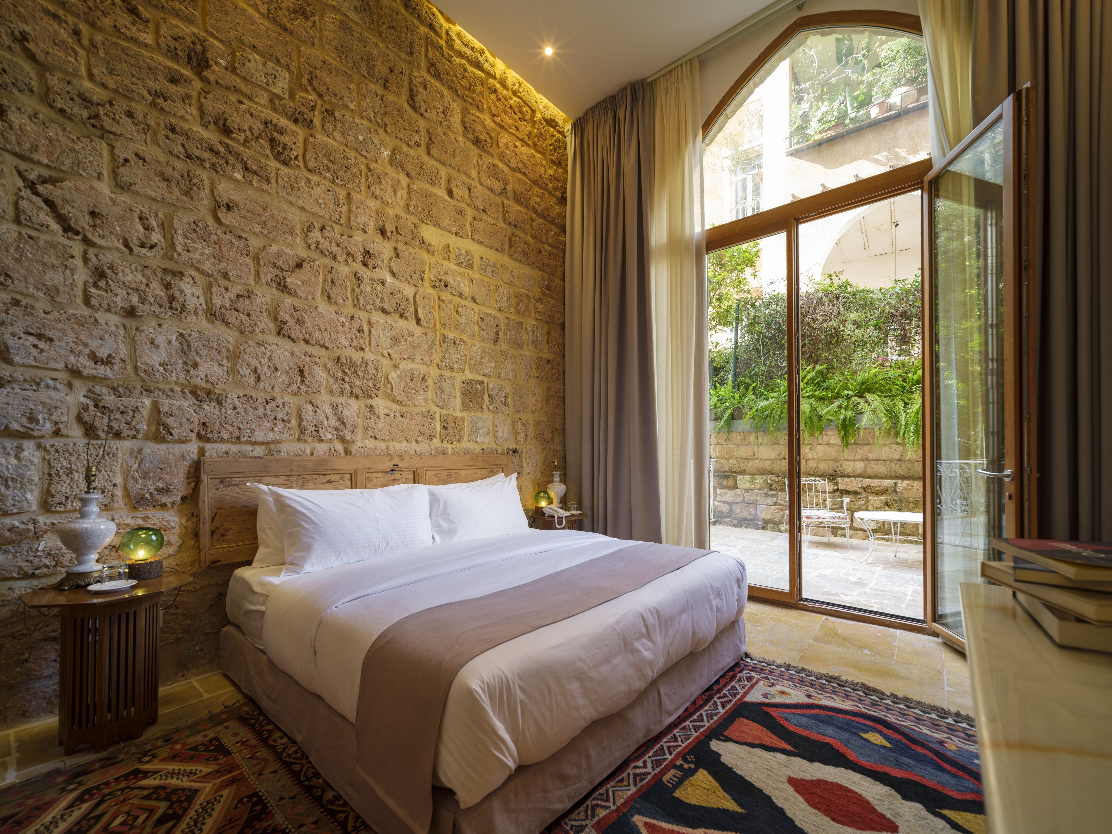 A bedroom with a stone wall, a bed with white linens, a patterned rug, and a view of greenery through a glass door.