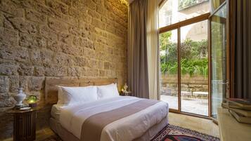 A bedroom with a stone wall, a bed with white linens, a patterned rug, and a view of greenery through a glass door.