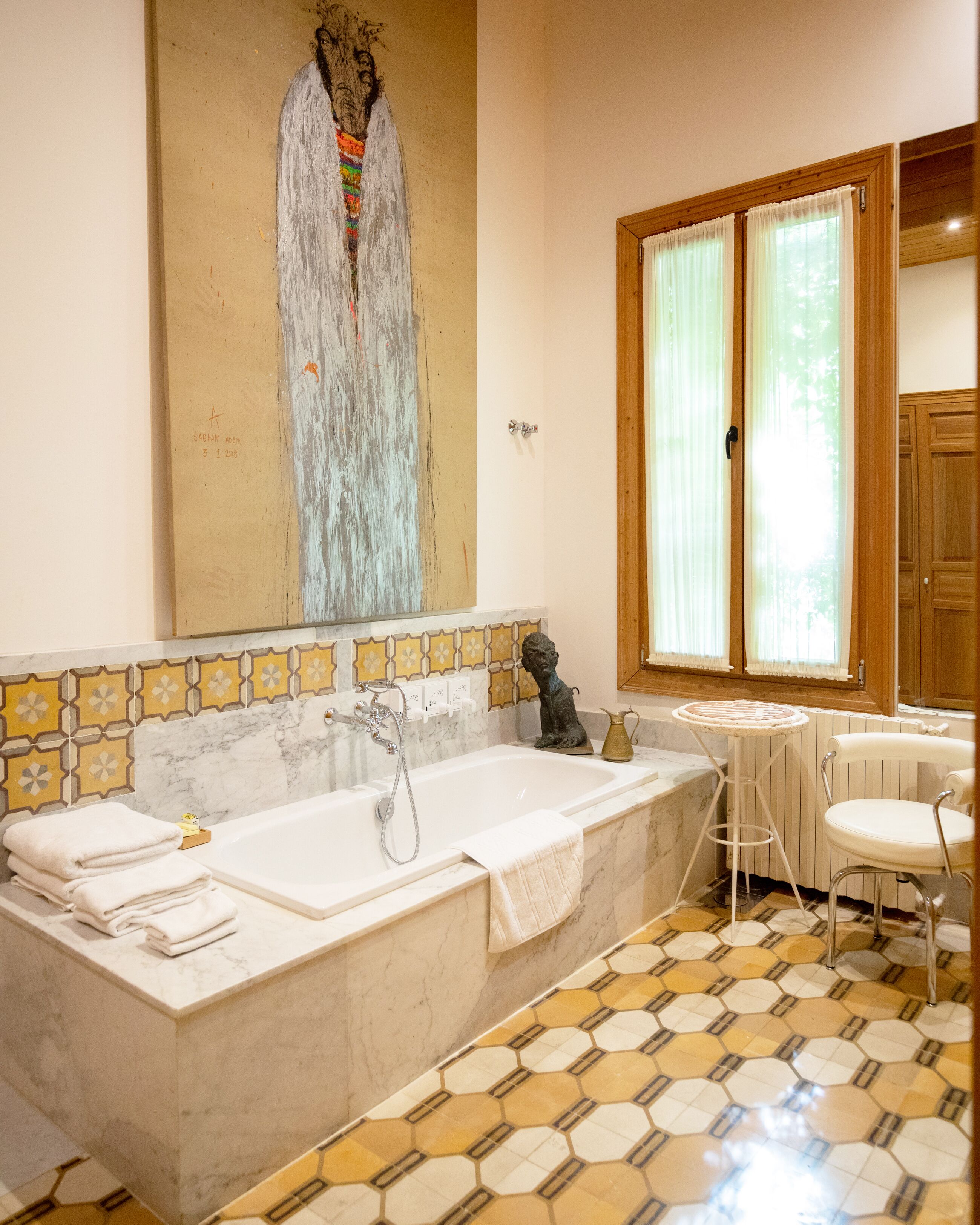 Un baño con una bañera grande, una mesita redonda pequeña, una silla y una ventana con cortinas.