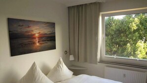 A bedroom with a bed, two pillows, a nightstand, a lamp, a window with a view of trees, and a painting of a sunset. - Rimmler - Südstrandperle (Sylt)