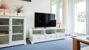 A modern living room with a flat-screen TV on a white console, a potted plant, and a lamp. - Rimmler - Südstrandperle (Sylt)