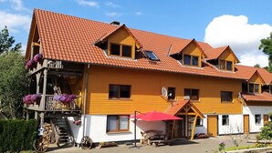 A multi-story wooden house with a red-tiled roof, a balcony with flower boxes, and a small attached structure with a red umbrella.
