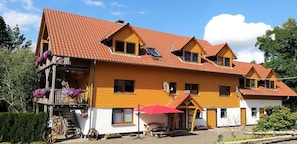 Una casa de madera de varios pisos con techo de tejas rojas, un balcón con macetas de flores y una pequeña estructura adosada con un paraguas rojo.