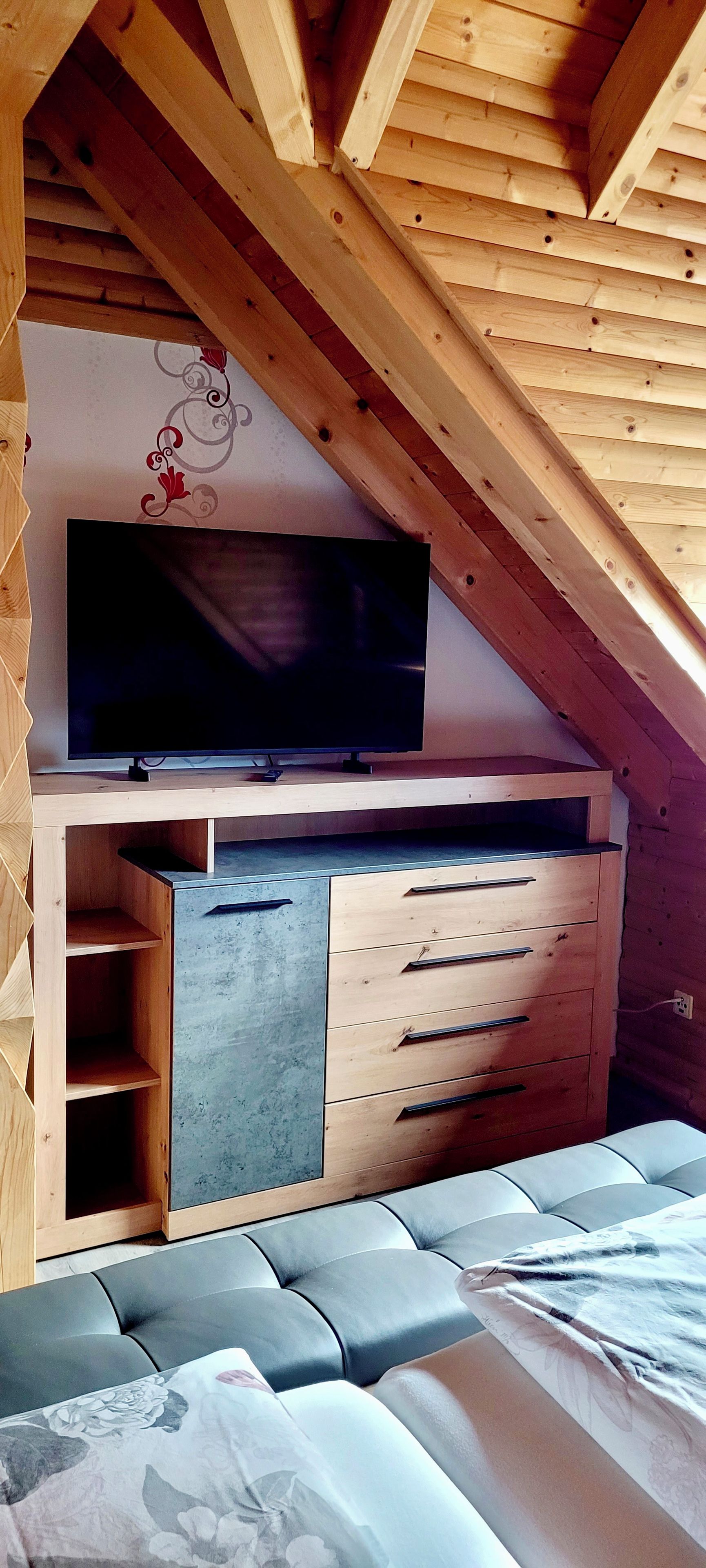 A bedroom with a sloped ceiling, a flat-screen TV mounted on the wall, a wooden dresser with drawers, and a bed with a floral-patterned cover.