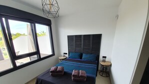 Een slaapkamer met een groot raam, een bed met blauwe en zwarte beddengoed, twee nachtkastjes en een geometrisch lamp.