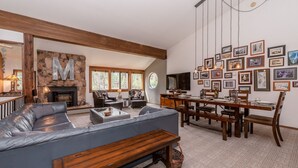 A cozy living room with a stone fireplace, a large gray sofa, wooden dining table, and a wall adorned with framed pictures. - Unique Alpine Condo! Steps away from Village Gondola! (Mammoth Lakes)