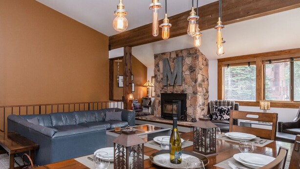 A cozy living room with a stone fireplace, a large 'M' on the wall, and a dining area set for four. - Unique Alpine Condo! Steps away from Village Gondola! (Mammoth Lakes)