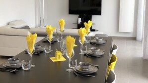 Una mesa de comedor con servilletas amarillas, vasos transparentes y platos grises en un salón moderno con televisor de pantalla plana y un sofá blanco.
