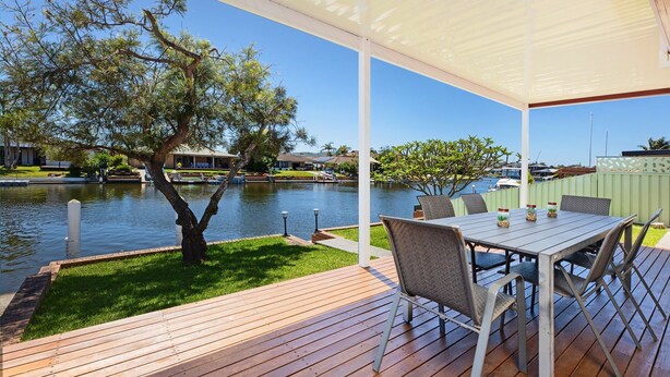 Una terraza de madera con una mesa y sillas, con vista a un cuerpo de agua con barcos y casas al fondo.