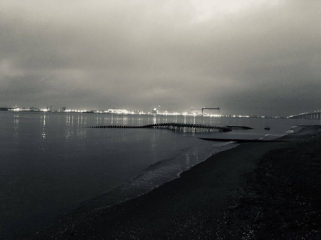 A nighttime coastal scene with a calm sea, a pier extending into the water, and distant city lights reflecting on the water's surface.