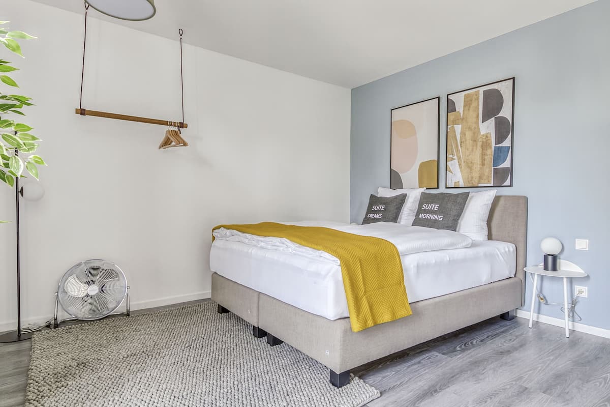 Une chambre moderne avec un lit, une table de chevet, un ventilateur sur pied et des œuvres d’art abstraites accrochées au mur.