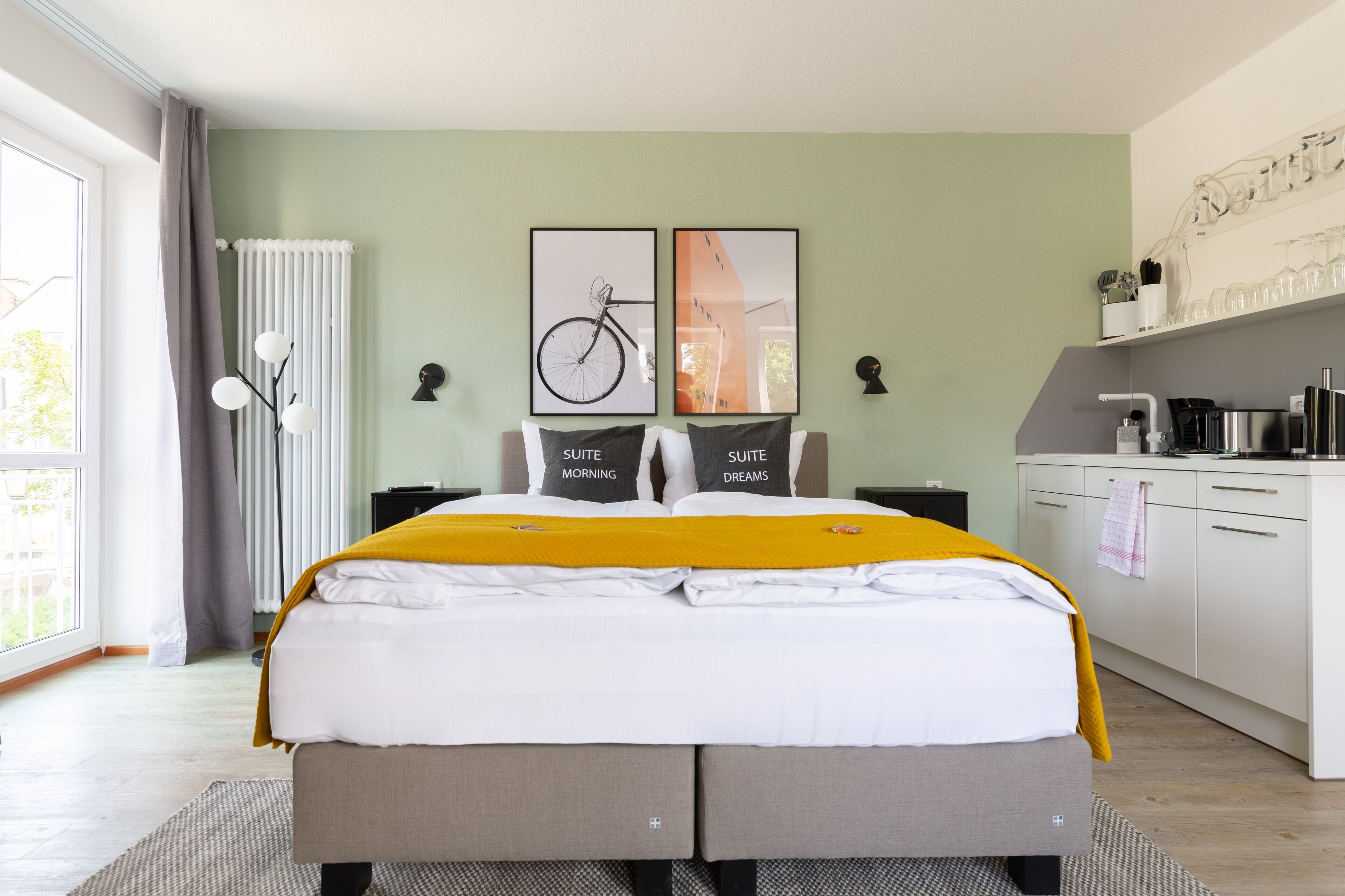 A modern hotel room with a bed, a kitchen area, and a large window.