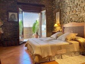A bedroom with a stone wall, a bed with a quilt, a nightstand with a lamp, and a window with blinds. - CAL SERRA - Country House (Alentorn)
