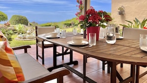 A round wooden table set for dining with a view of the ocean, surrounded by chairs and a potted plant with red flowers. - Andalusian style villa with stunning views. PAX 6. FREE WIFI (Tarifa)