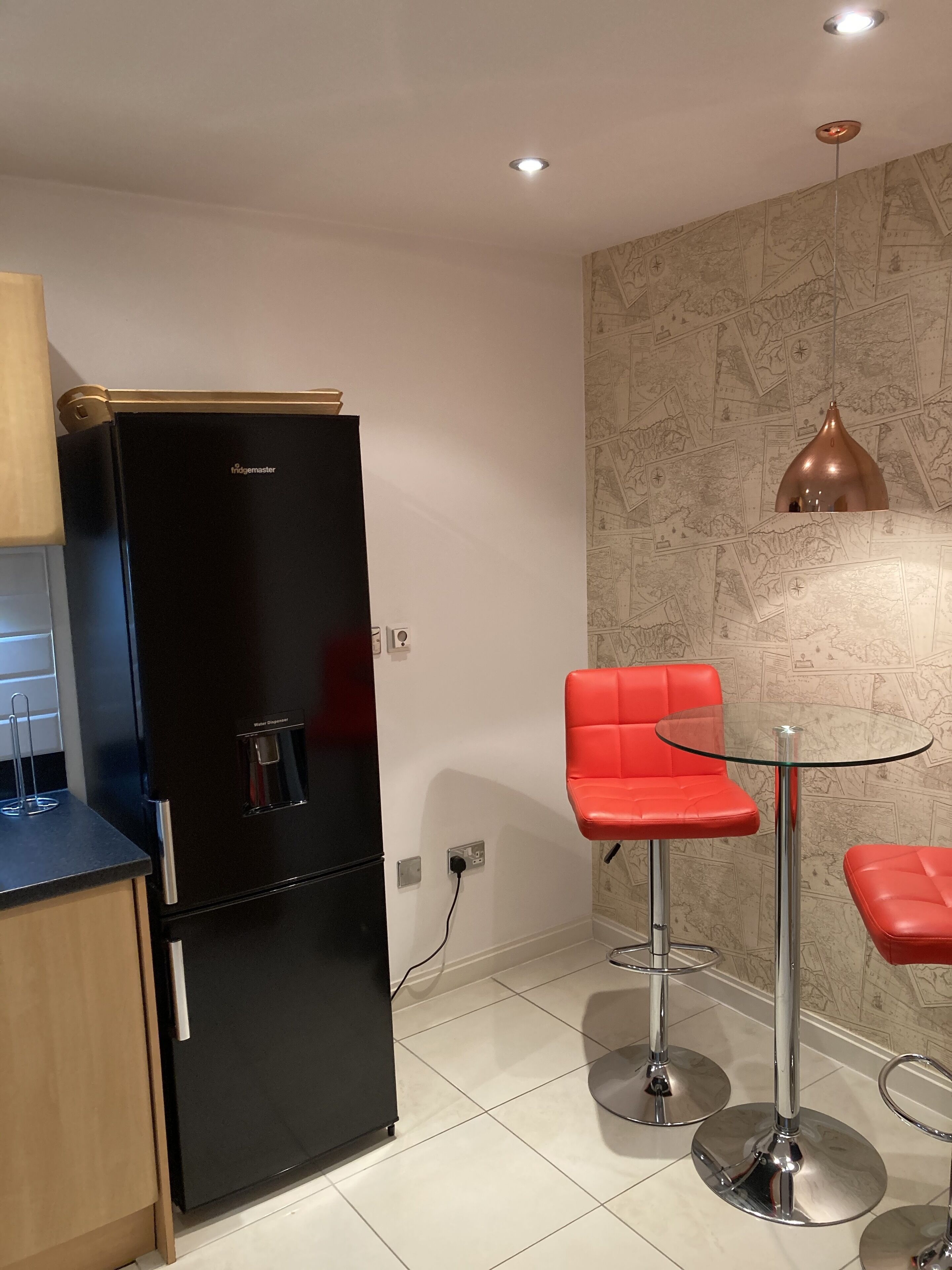 A modern kitchen with a black refrigerator, a water dispenser, and a red bar stool with a glass table.