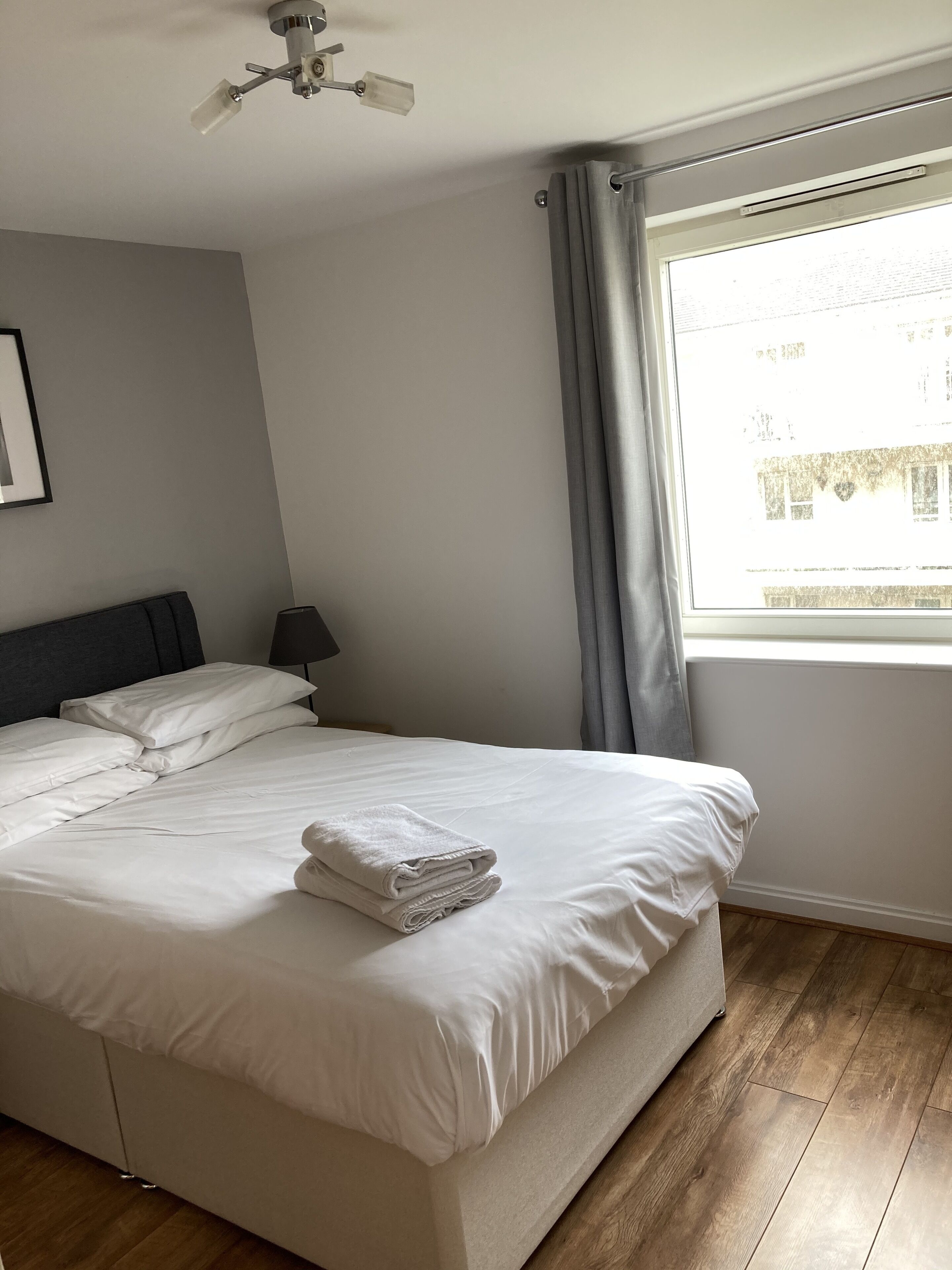 Una cama bien hecha con sábanas blancas y una toalla doblada, un ventilador de techo, una mesita de noche con lámpara y una ventana con cortinas.