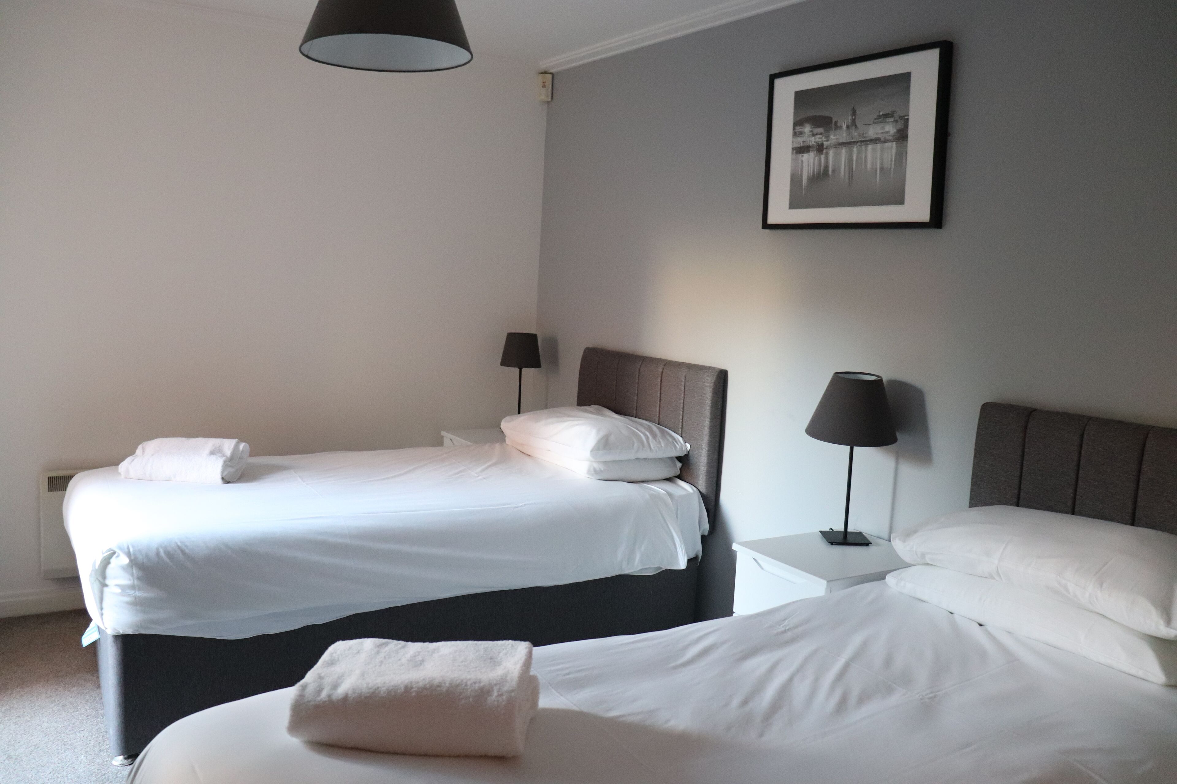 A hotel room with two beds, each with white linens and pillows, a bedside table with a lamp, and a framed picture on the wall.