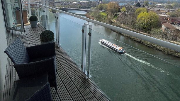 A balcony with a view of a river and a boat. There are wicker chairs and a potted plant.