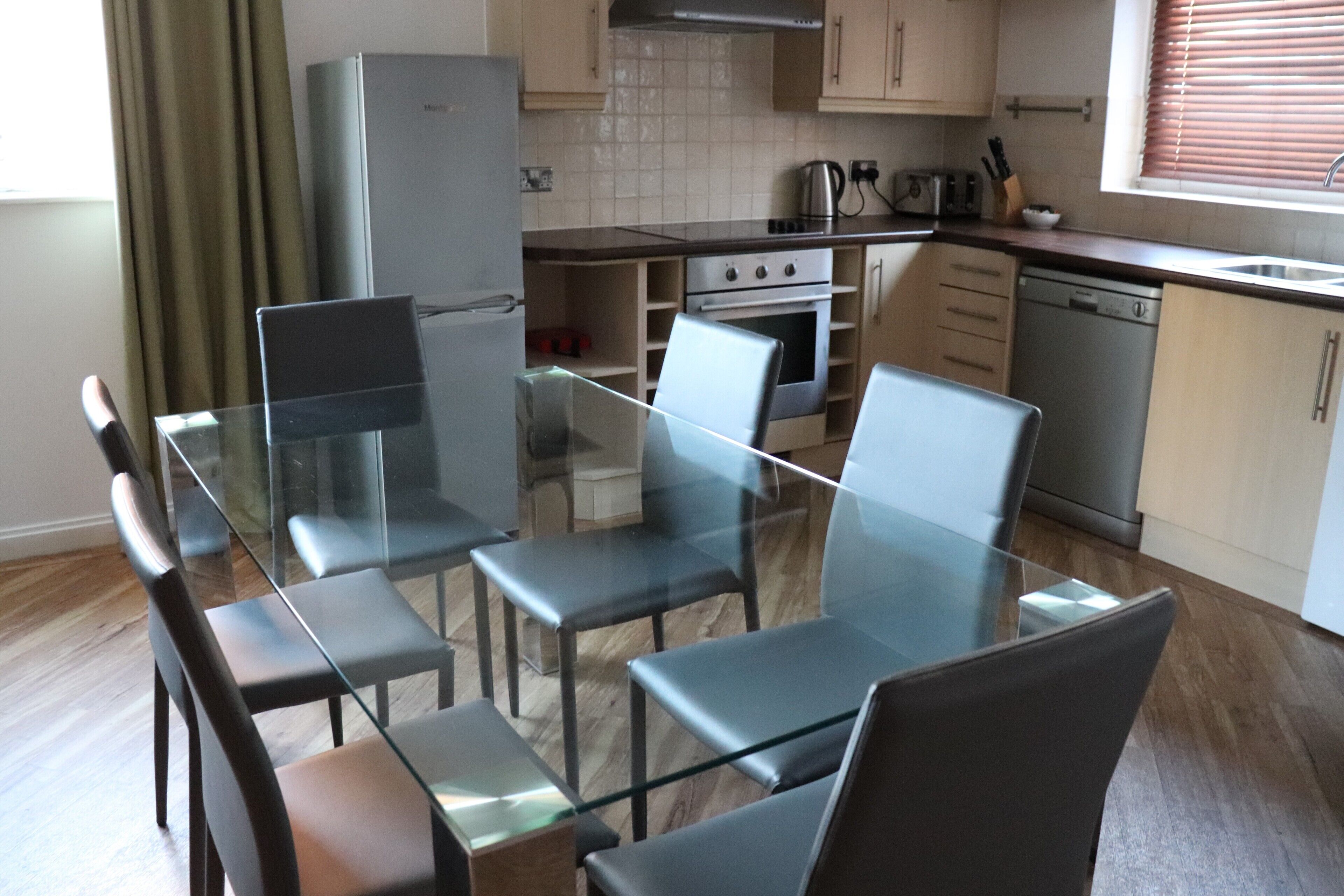 A modern kitchen with a glass dining table and chairs, wooden cabinets, a built-in oven, and a dishwasher.